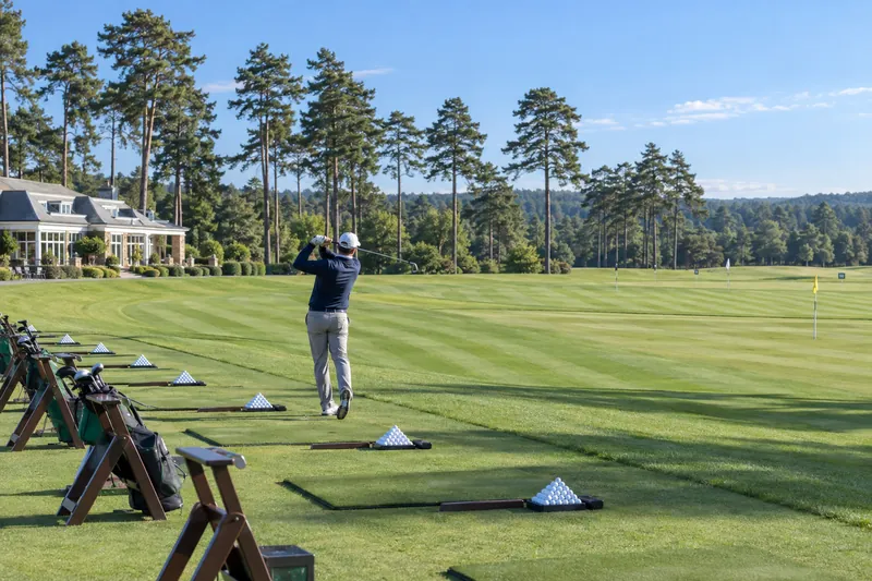 A visitor hits balls on the practice ground in the sun, the clubhouse and a line of Scots pines behind.